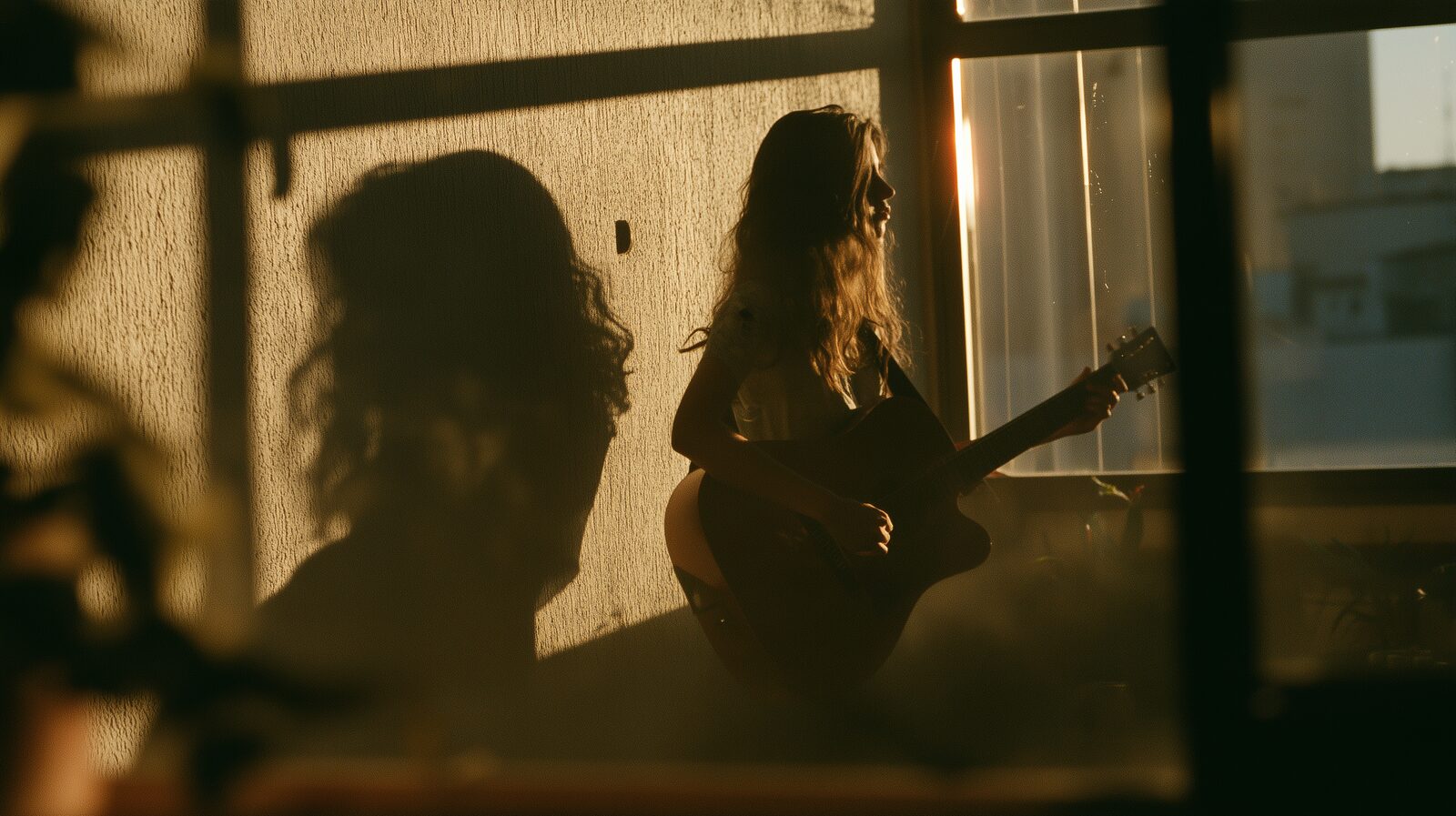 Julia toca violão contra a luz do nascer do sol, junto à janela de um apartamento em São Paulo, e a sombra dela se projeta grande na parede. O rosto fica na sombra