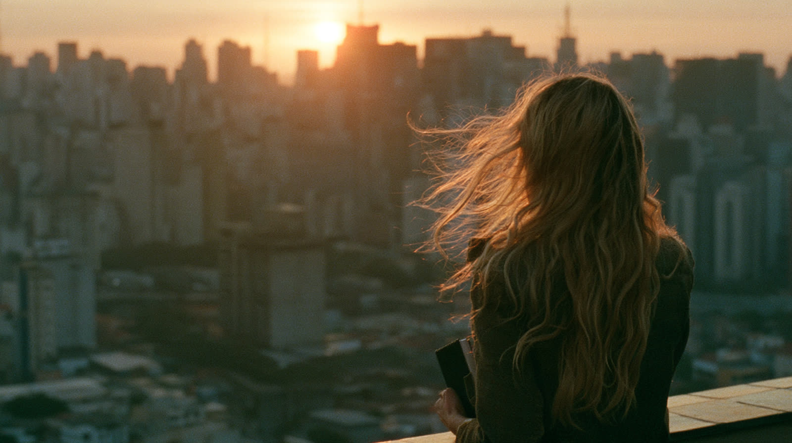 Julia, de costas, com o cabelo loiro ao vento, olha São Paulo do alto de um prédio ao nascer do sol. O rosto não aparece