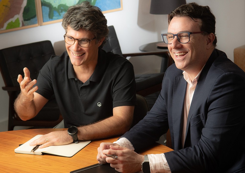 Daniel Lemos, da Riza Asset, e Paulo Barcellos, da O2 Filmes, conversam sorrindo à mesa