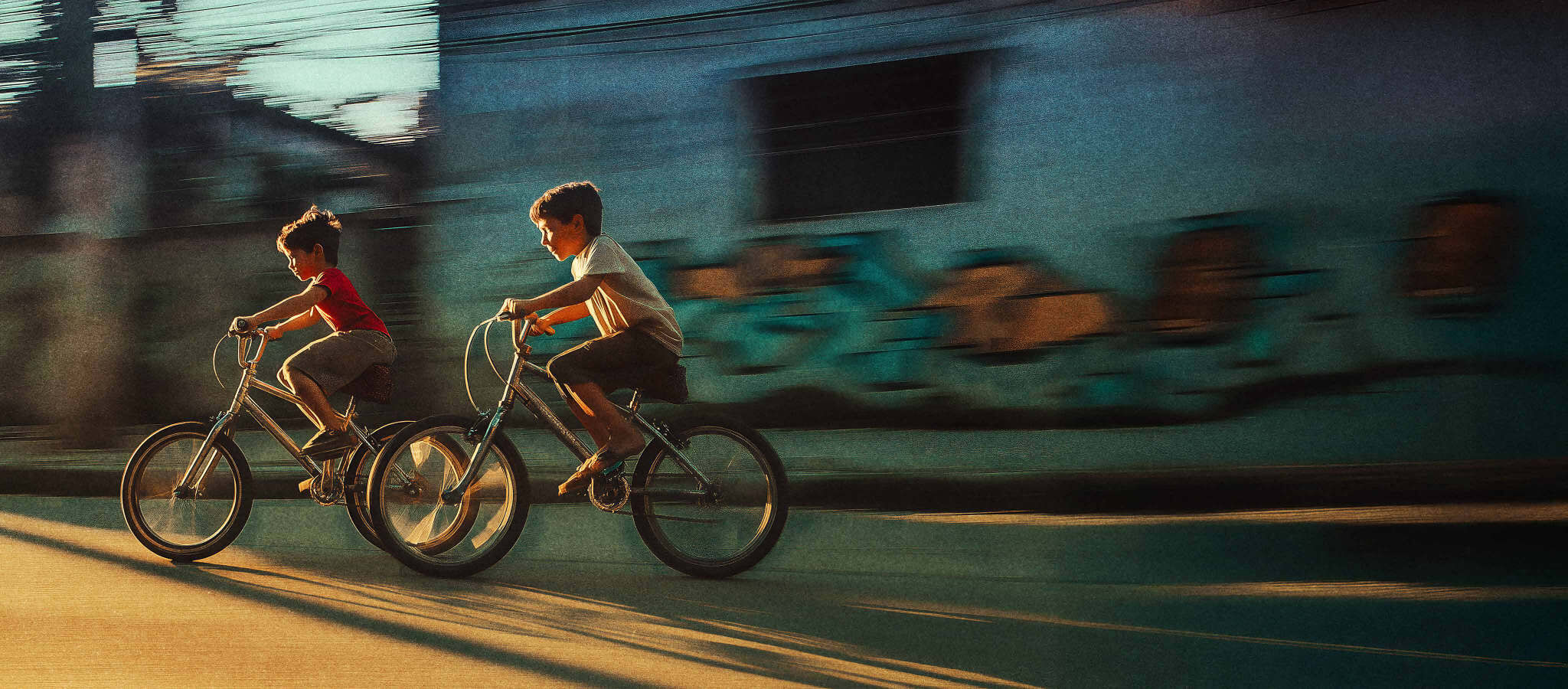 Dois meninos pedalam lado a lado numa rua de periferia, com borrão de velocidade