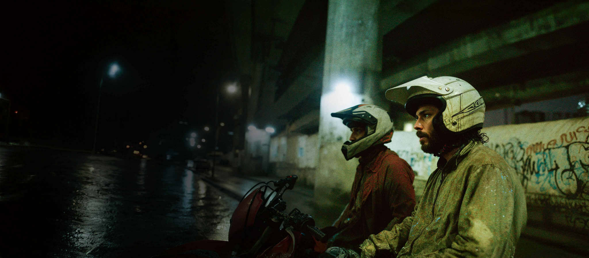 Dois motociclistas de capacete sob um viaduto, à noite, na chuva