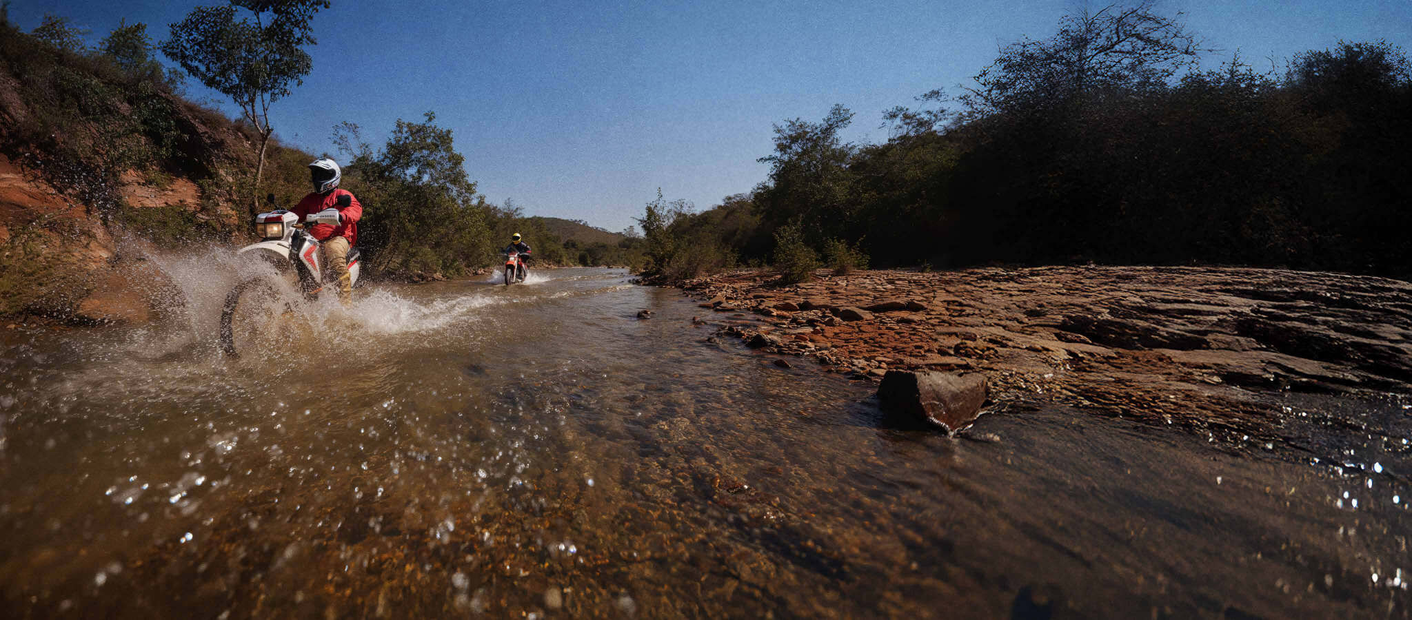 Moto atravessa um rio raso espirrando água, sob céu azul