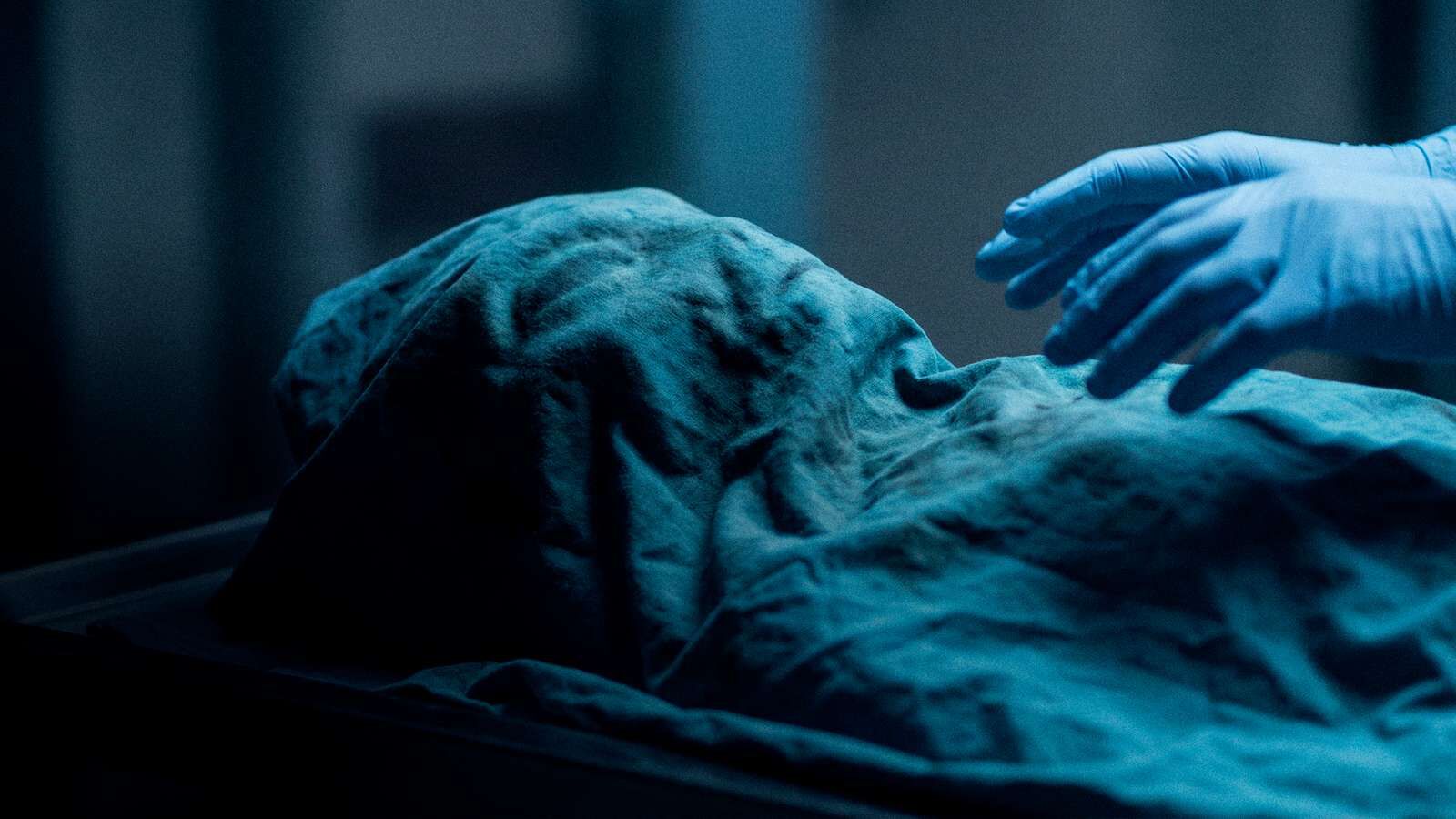Mão com luva azul sobre um corpo coberto por lençol, em luz fria